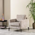 Een beige gestoffeerde fauteuil met donkere houten poten staat naast een zwarte ronde bijzettafel met een vaas met bloemen en kleurrijke kaarsen. Een potplant staat ernaast. De kamer heeft lichtgekleurde muren en een betegelde vloer.