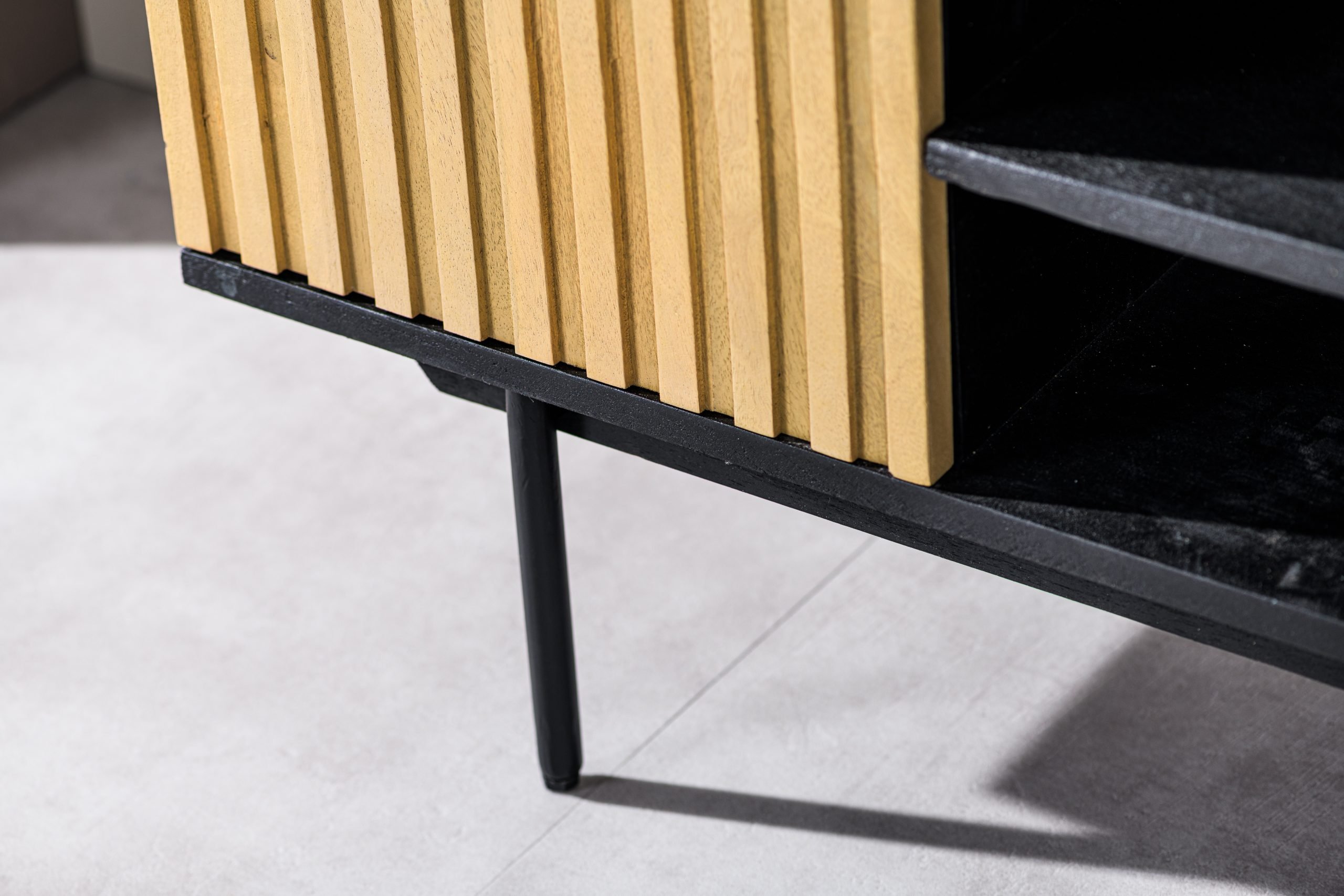 Close-up van een moderne kast met verticale houten latten en zwarte metalen poten. Het contrasterende lichte hout en donkere metaal creëren een strakke, eigentijdse look, met een deel van de planken zichtbaar aan de rechterkant. De vloer is lichtgrijs.