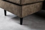 Close-up van de hoek van een bruine, suède-achtige gestoffeerde bank met zwarte houten poten, zittend op een gladde grijze vloer. De afbeelding richt zich op de textuur en details van de stof en het strakke, moderne ontwerp van de poten.