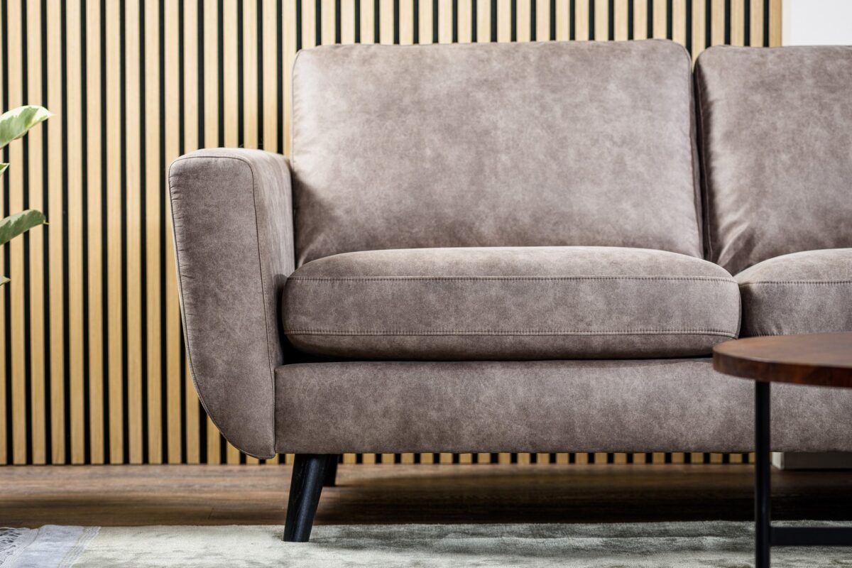 Een close-up van de 3 zits bank CL L+R in Savannah S520 taupe stof, met zwarte poten op een licht vloerkleed voor een verticale houten lattenwand, met glimpen van een plant en een deel van een ronde houten tafel.