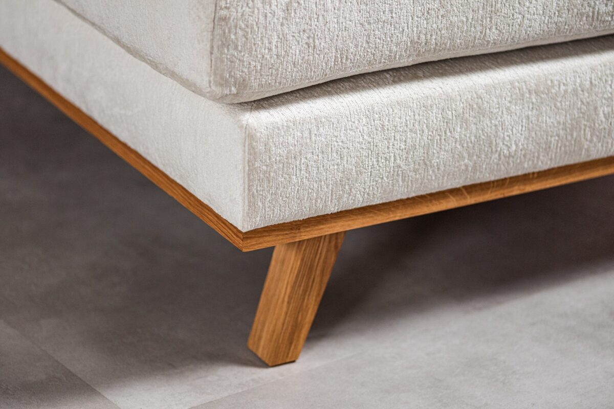 Close-up van een hoek van een lichtbeige bank met houten poten. De textuur van de stof is zichtbaar en de bank staat tegen een lichtgrijze tegelvloer. De houten basis en poten contrasteren met de zachte bekleding.