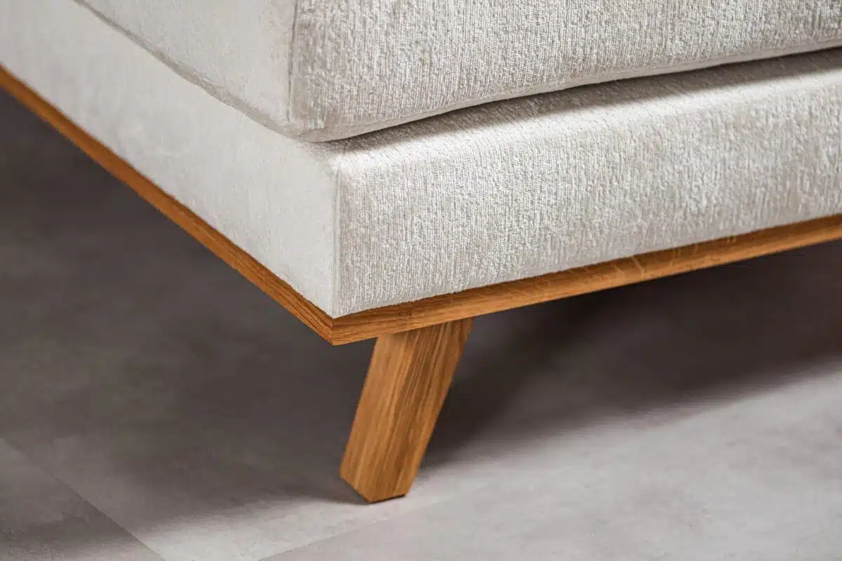 Close-up van een hoek van een lichtbeige bank met houten poten. De textuur van de stof is zichtbaar en de bank staat tegen een lichtgrijze tegelvloer. De houten basis en poten contrasteren met de zachte bekleding.