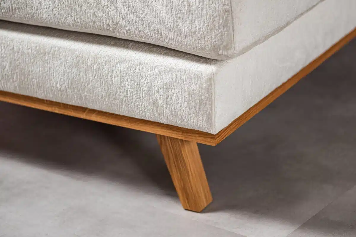 Close-up van een lichtgrijze gestoffeerde hoekbank met houten poten, geplaatst op een gladde, lichtgekleurde vloer. Het ontwerp heeft een minimalistische en moderne esthetiek met een focus op de hoekige houten pootondersteuning.