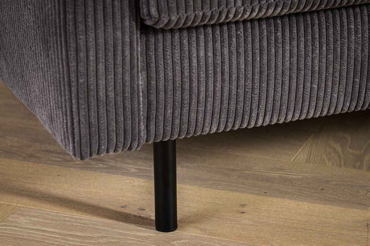 Close-up van de hoek van een grijze corduroy bank met geribbelde textuur. De bank heeft een zwarte metalen poot en rust op een houten vloer met een lichtbruine afwerking.