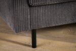 Close-up van de hoek van een grijze corduroy bank met geribbelde textuur. De bank heeft een zwarte metalen poot en rust op een houten vloer met een lichtbruine afwerking.