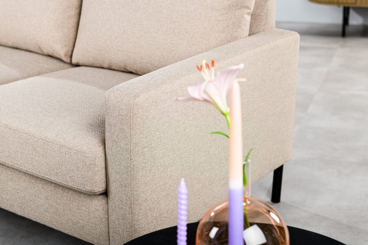 Een beige bank op een lichtgekleurde vloer met een klein zwart tafeltje ervoor. Op het tafeltje staat een roze vaas met een witte lelie en drie gedraaide kaarsen in pastelkleuren: paars, roze en perzik. Op de achtergrond is een deel van een houten kast te zien.