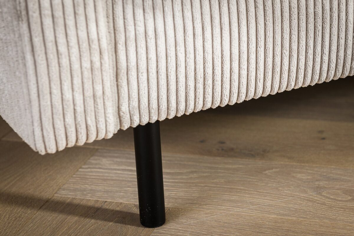 Close-up van een lichtbeige corduroy-textuur bank arm of zijkant. Een zwarte cilindrische poot ondersteunt het op een lichte houten vloer. De textuur en materialen zijn duidelijk zichtbaar, waardoor de geribbelde details van de stof worden benadrukt.