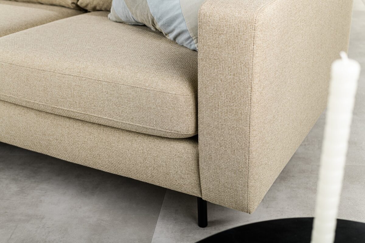 Close-up van een beige bank in een moderne woonkamer. De stof heeft een subtiel structuurpatroon. Een kussen met een gestreept design is gedeeltelijk zichtbaar. Een witte kaars staat op een zwart oppervlak op de voorgrond.