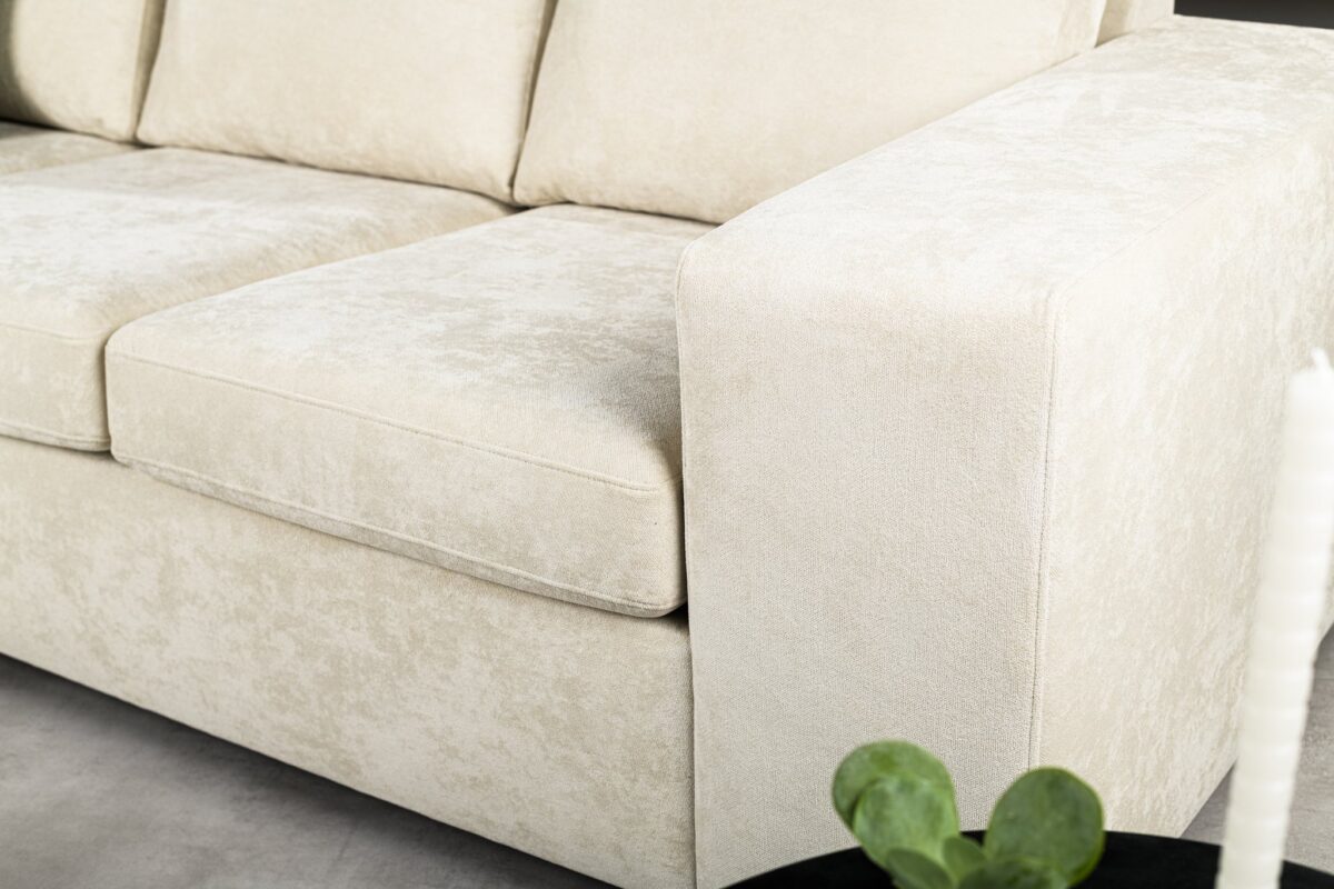 Beige gestoffeerde bank met pluche kussens en brede armleuningen, geplaatst op een lichtgekleurde vloer. Een klein deel van een ronde, donkergekleurde tafel met een plant is zichtbaar op de voorgrond.