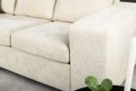 Beige gestoffeerde bank met pluche kussens en brede armleuningen, geplaatst op een lichtgekleurde vloer. Een klein deel van een ronde, donkergekleurde tafel met een plant is zichtbaar op de voorgrond.