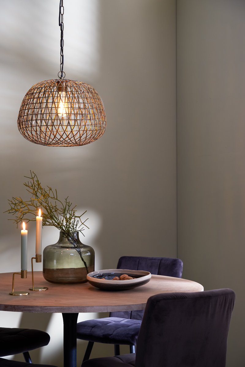 Een gezellige eethoek heeft een ronde houten tafel met een keramische kom en een vaas met takken. Er staan twee brandende kaarsen in de buurt. Erboven werpt een geweven, bolvormige hanglamp een warme gloed. Donkere gestoffeerde stoelen omringen de tafel.