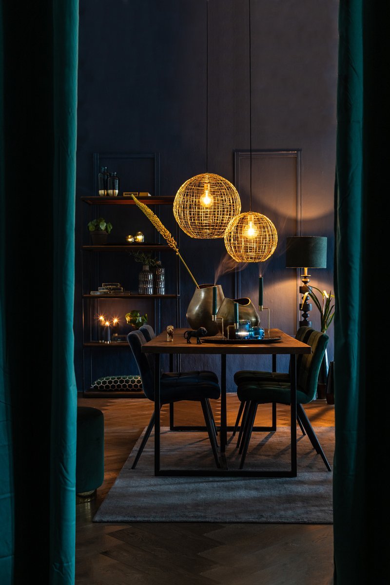 Een schemerige eetkamer met een houten tafel en donkere stoelen. Twee bolvormige hanglampen hangen boven de tafel en werpen een warme gloed. Planken met decoratie en planten staan op de achtergrond, samen met een staande lamp. Gordijnen omlijsten het tafereel.