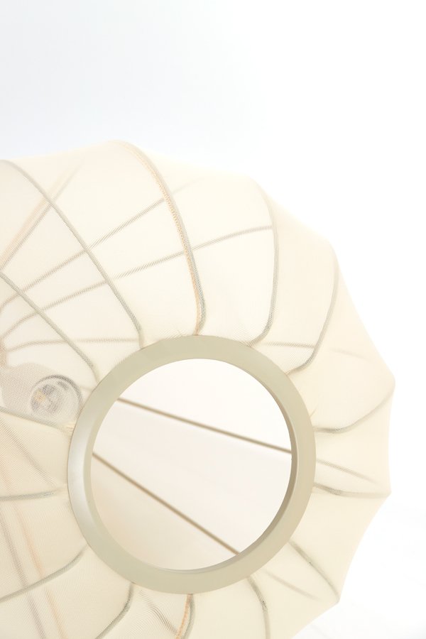 Close-up van een moderne, ronde lampenkap met een lichtbeige stof en een lichtmetalen frame. De stof is gespannen over een draadstructuur, waardoor een geometrisch, artistiek ontwerp ontstaat tegen een witte achtergrond.