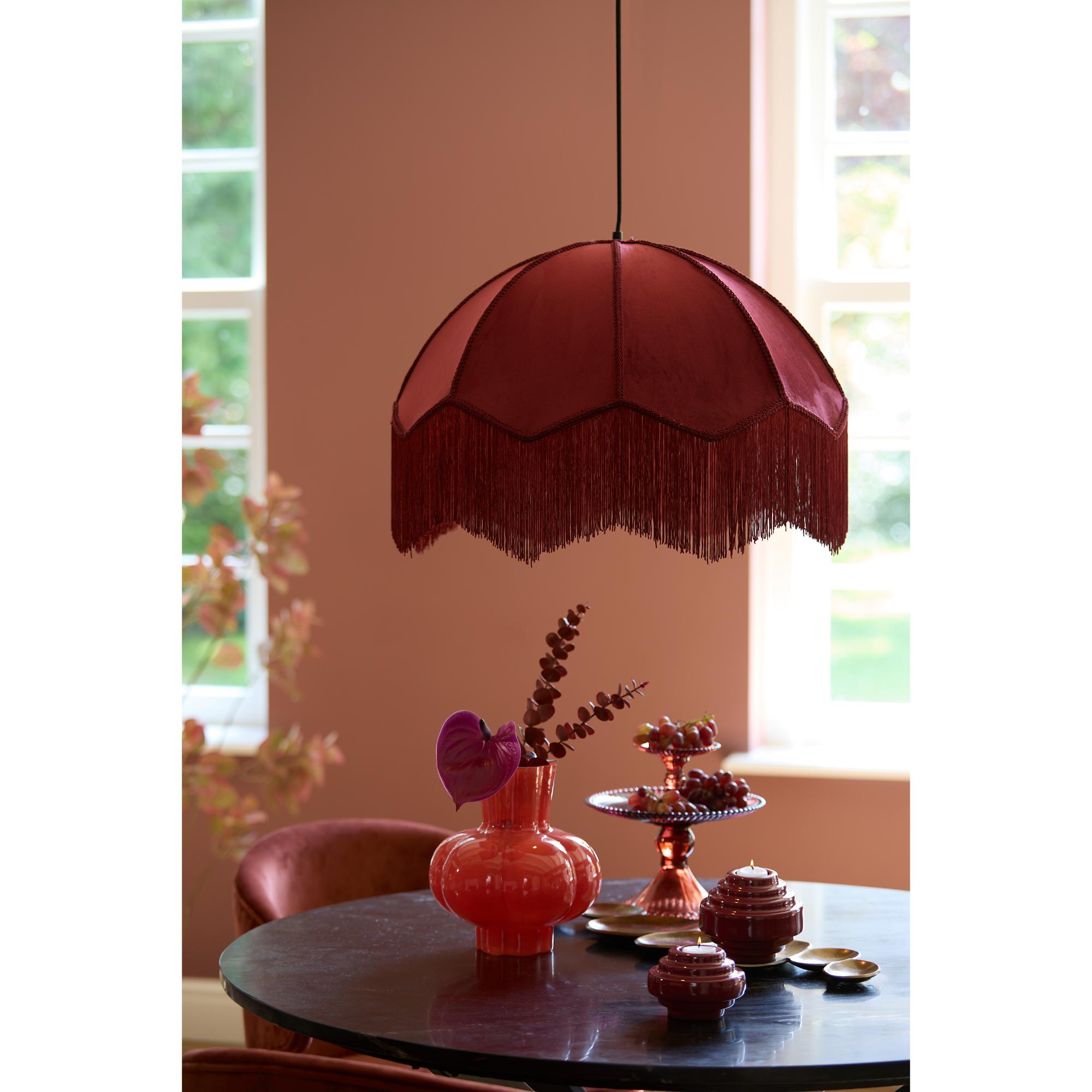 Een gezellige eethoek heeft een ronde tafel met decoratieve rode glazen objecten, waaronder vazen en snoepschalen. Een grote, gefranjerde kastanjebruine lampenkap hangt erboven. Zacht licht filtert door nabijgelegen ramen, wat een warme ambiance creëert.