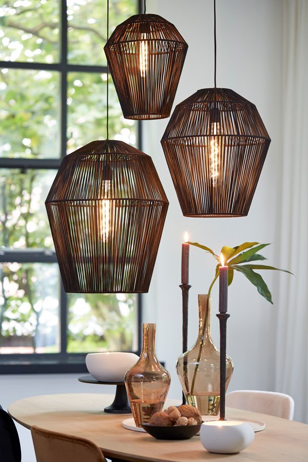 Drie geometrische hanglampen hangen boven een eettafel met decoratieve glazen flessen, brandende kaarsen en een schaal met noten. Een groot raam op de achtergrond laat natuurlijk licht binnen, wat een gezellige ambiance creëert.