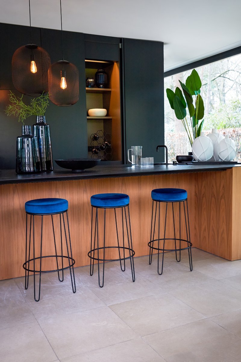 Moderne keuken met een houten eiland met drie blauwe fluwelen barkrukken met zwarte metalen poten. Boven het eiland hangen twee hanglampen. Planken met decoratie-items zijn ingebouwd in een donkere muur en grote ramen laten een tuin buiten zien.