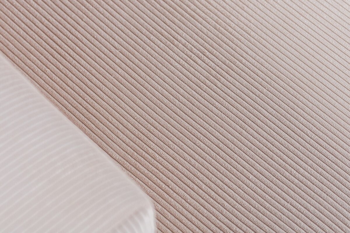 Close-up van een gestructureerd oppervlak met geribbelde strepen in een lichtbeige kleur. Een klein, gevouwen stukje witte stof met dunne, parallelle lijnen is zichtbaar in de hoek, gedeeltelijk overlappend met het beige materiaal.