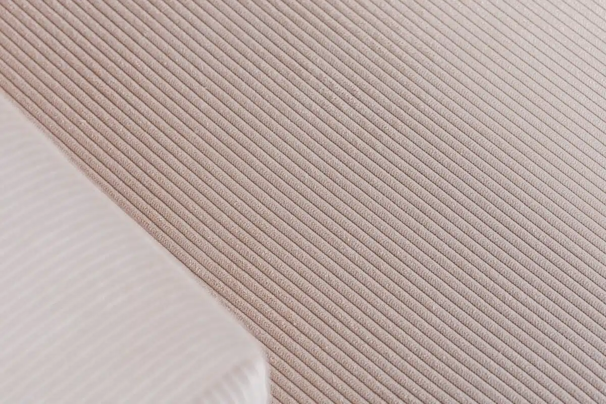 Close-up van een gestructureerd oppervlak met geribbelde strepen in een lichtbeige kleur. Een klein, gevouwen stukje witte stof met dunne, parallelle lijnen is zichtbaar in de hoek, gedeeltelijk overlappend met het beige materiaal.