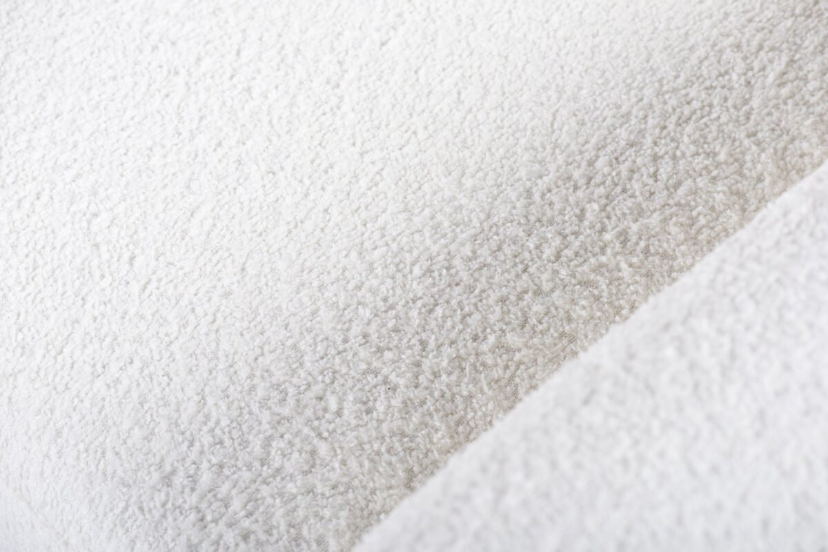 Close-up van witte stof met een zacht, gestructureerd oppervlak, dat lijkt op fleece of sherpa-materiaal. De afbeelding toont een diagonale naad, die diepte toevoegt aan het pluche uiterlijk.