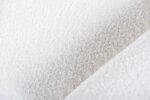 Close-up van witte stof met een zacht, gestructureerd oppervlak, dat lijkt op fleece of sherpa-materiaal. De afbeelding toont een diagonale naad, die diepte toevoegt aan het pluche uiterlijk.
