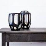 Twee moderne zwarte lantaarns met geometrische uitsparingen staan op een rustieke houten tafel tegen een effen witte achtergrond. De lantaarns hebben een strak, eigentijds design en zijn dicht bij elkaar geplaatst.