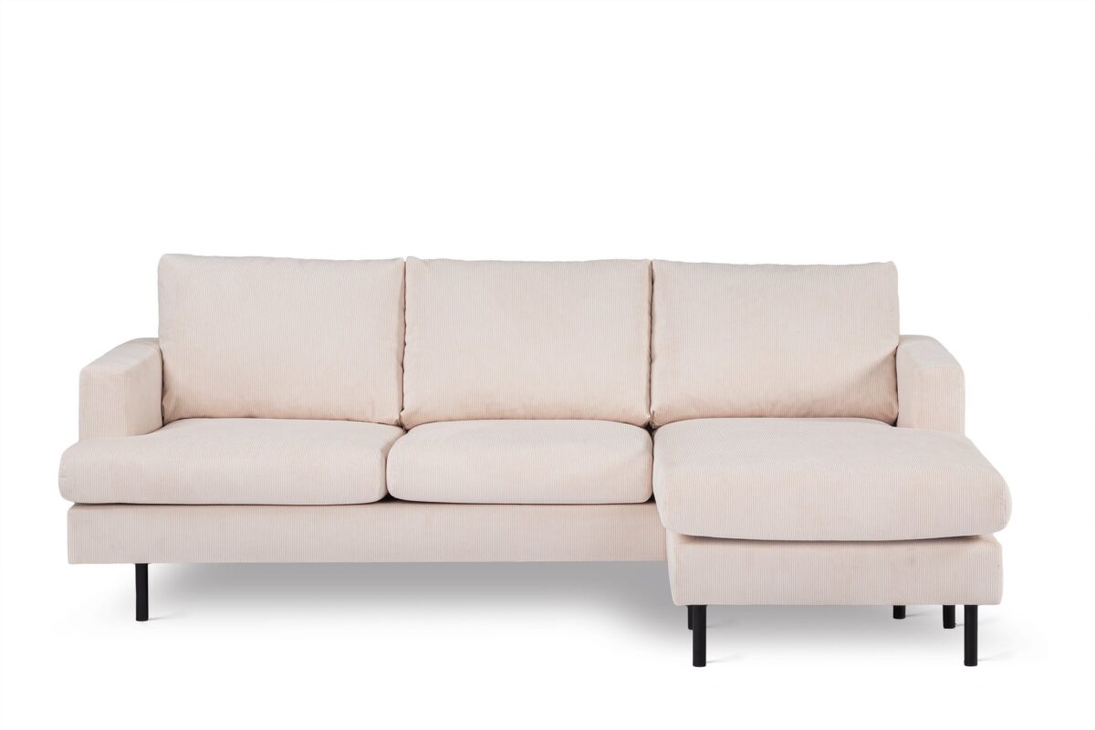 Een crèmekleurige L-vormige sectionele bank met drie zitkussens en een chaise longue aan de rechterkant. Het heeft zwarte poten en een minimalistisch design, tegen een effen witte achtergrond.