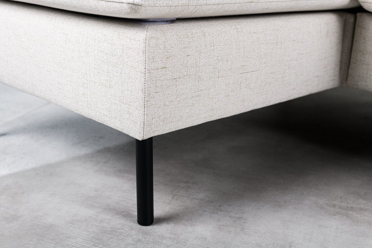 Close-up van een lichtbeige stoffen hoekbank met minimalistisch design. De bank heeft een zwarte metalen poot en rust op een grijze vloer met textuur. De bekleding heeft een subtiel geweven patroon.