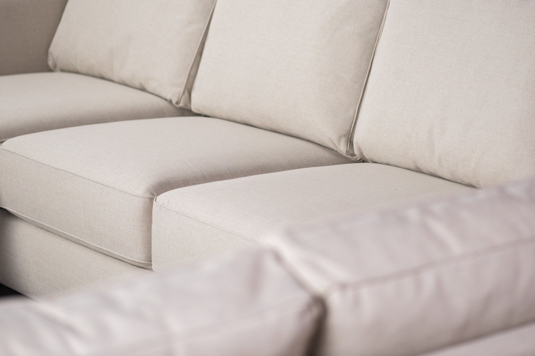 Close-up van een beige stoffen bank met pluche kussens. De focus ligt op de zitting en de rugleuning, waarbij de textuur en het stikseldetail zichtbaar zijn. De hoek van de bank is zichtbaar op de voorgrond.