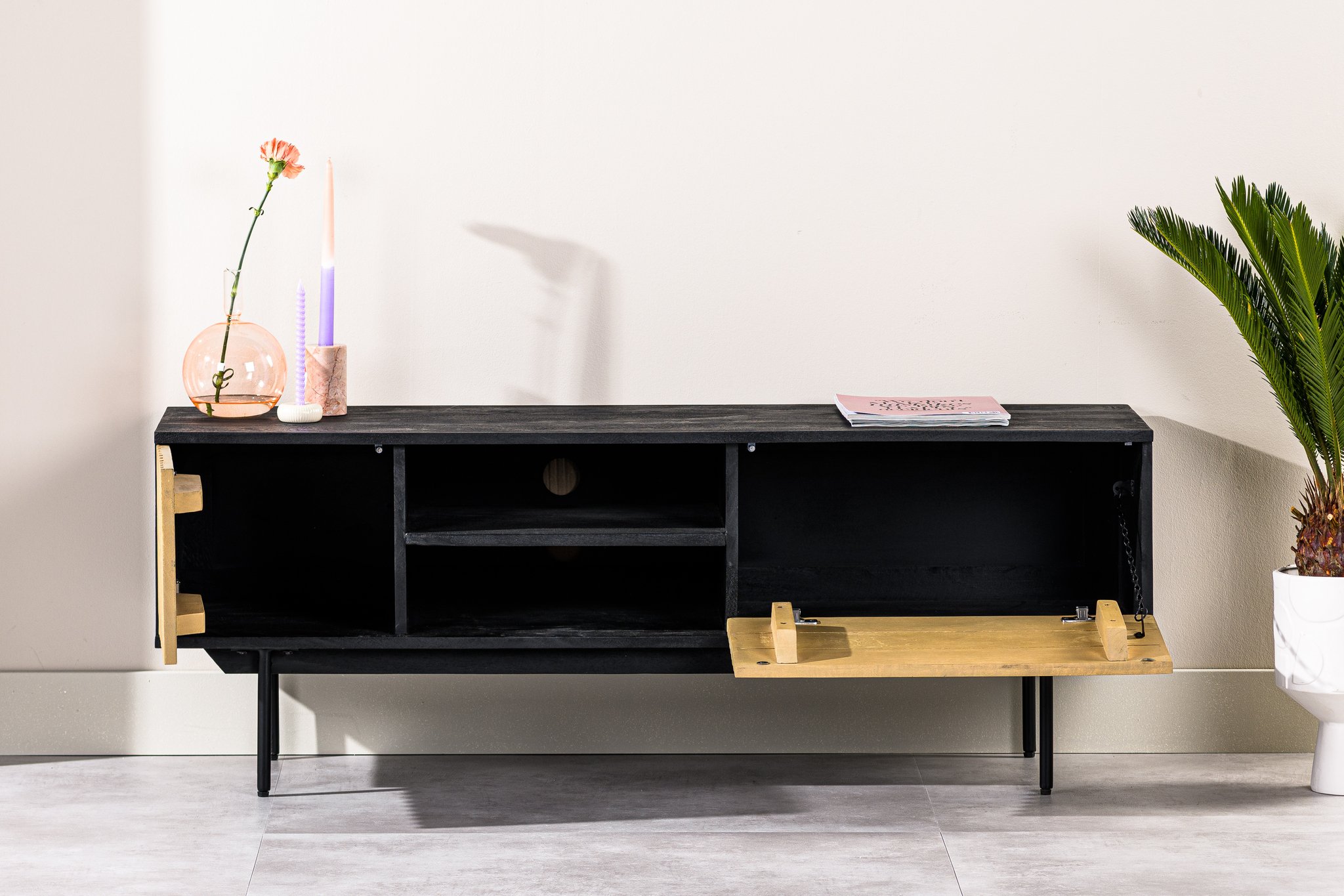 Een moderne zwarte tv-standaard met open vakken en een houten klapdeur aan de rechterkant staat tegen een crèmekleurige muur. Er staan een kaars, een bloem in een vaas en een dicht boek op. Rechts staat een potplant met brede groene bladeren.