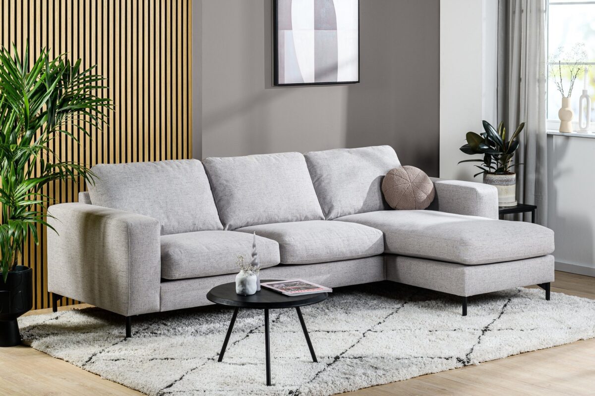 Een moderne woonkamer met de 3 zits bank CL L+R in Valente V311 grijs, gecombineerd met een ronde zwarte salontafel, geometrisch vloerkleed, hoge groene plant en muurkunst tegen houten latten en een grijs geschilderde muur.