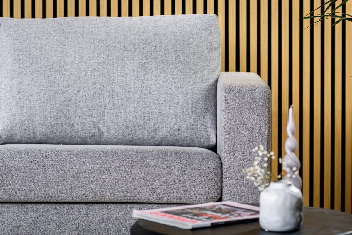 De 3-zits bank CL L+R in Chloe C311 grijs stof staat voor een verticale houten lattenwand. Op de voorgrond staat een zwarte tafel met een kleine witte vaas, decoratieve stokken en een tijdschrift.