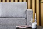 De 3-zits bank CL L+R in Chloe C311 grijs stof staat voor een verticale houten lattenwand. Op de voorgrond staat een zwarte tafel met een kleine witte vaas, decoratieve stokken en een tijdschrift.