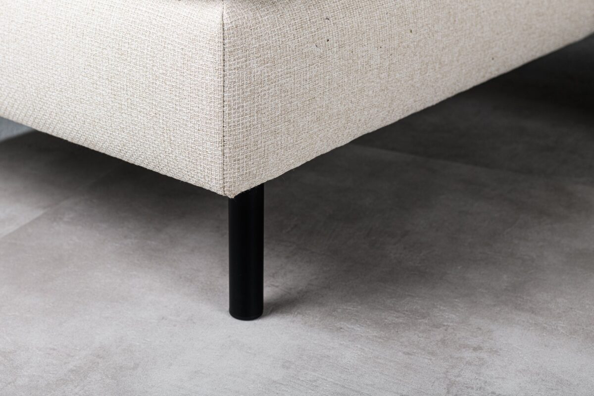 Close-up van de hoek van de 3 zits bank CL L+R in D820 ivoor stof Dillon, met zijn beige bekleding en eenvoudige zwarte cilindervormige poot, op een lichtgrijze tegelvloer.