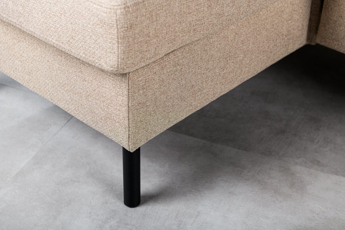 Close-up van een beige stoffen hoekbank met zwarte metalen poten, geplaatst op een lichtgrijze betegelde vloer. De textuur van de bank is grof en de bank is vanuit een lage hoek te zien.