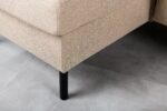Close-up van een beige stoffen hoekbank met zwarte metalen poten, geplaatst op een lichtgrijze betegelde vloer. De textuur van de bank is grof en de bank is vanuit een lage hoek te zien.