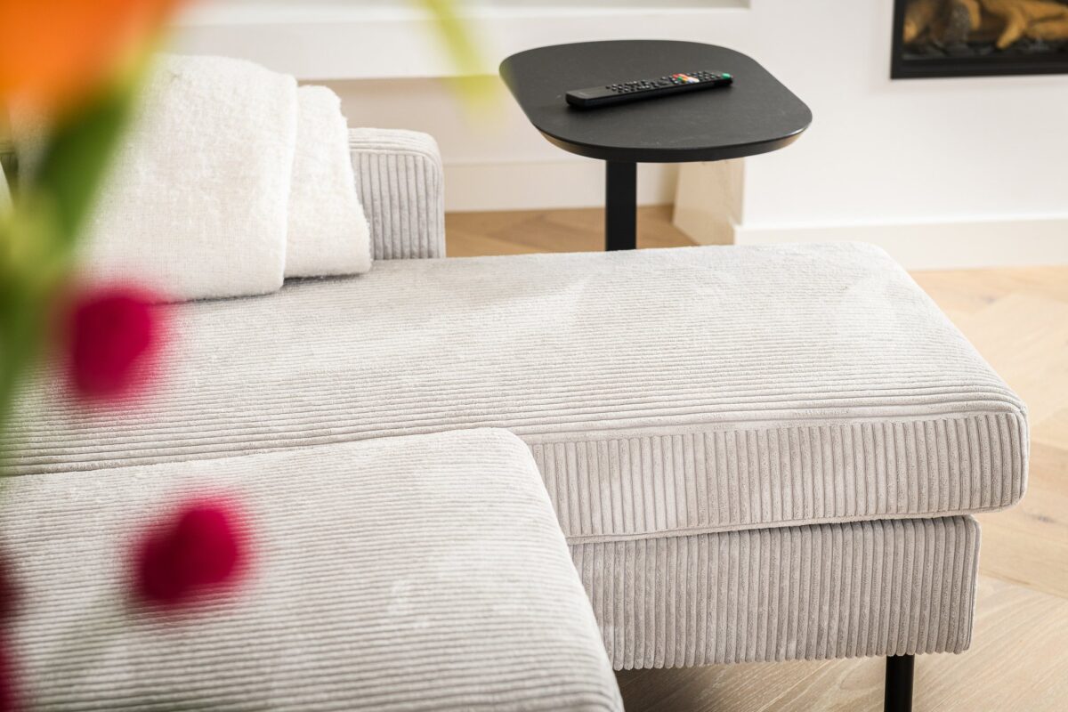 Een gezellige, lichtgrijze corduroy chaise longue met een witte deken eroverheen gedrapeerd staat naast een klein zwart bijzettafeltje met een afstandsbediening. Op de voorgrond zijn wazige rode en oranje bloemen te zien. Een open haard is gedeeltelijk zichtbaar op de muur.