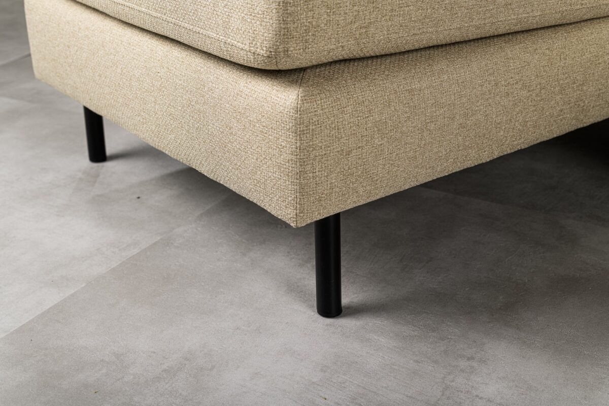 Close-up van een beige stoffen hoekbank met strakke zwarte metalen poten, geplaatst op een lichtgrijze betegelde vloer. De textuur van de stof van de bank is zichtbaar, wat het geweven patroon benadrukt.