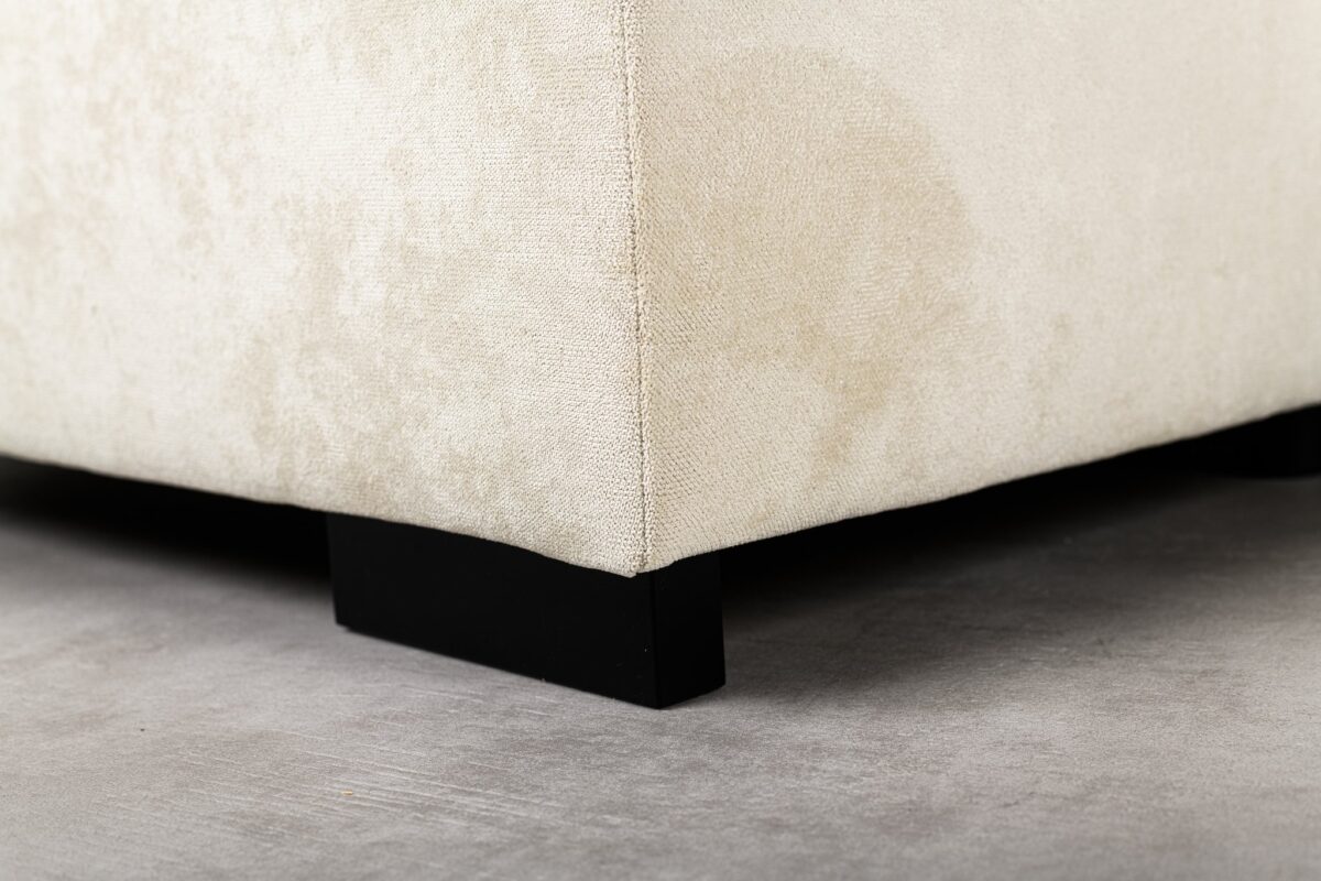 Close-up van de hoek van een beige gestoffeerde bank met een kleine vlek, ondersteund door zwarte houten poten op een gladde betonnen vloer.