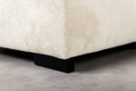 Close-up van de hoek van een beige gestoffeerde bank met een kleine vlek, ondersteund door zwarte houten poten op een gladde betonnen vloer.