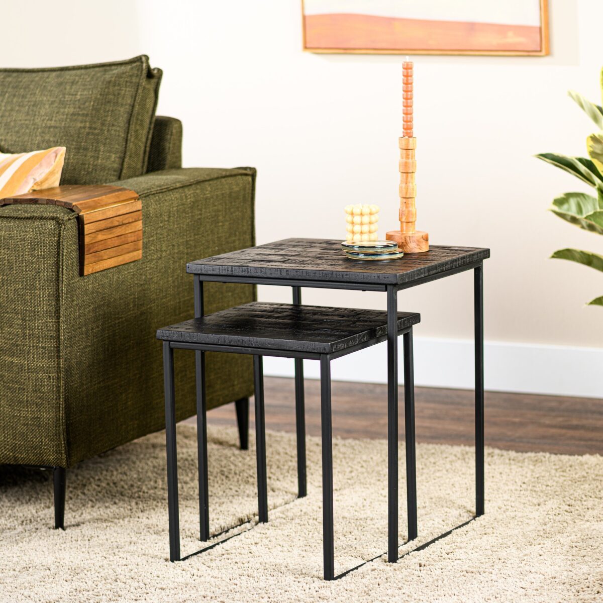 Twee moderne bijzettafels met zwarte metalen frames en houten bladen staan naast een groene bank. Een stapel houten blokken en een klein decoratief item staan op de grotere tafel. Een plant en een ingelijst kunstwerk staan op de achtergrond.