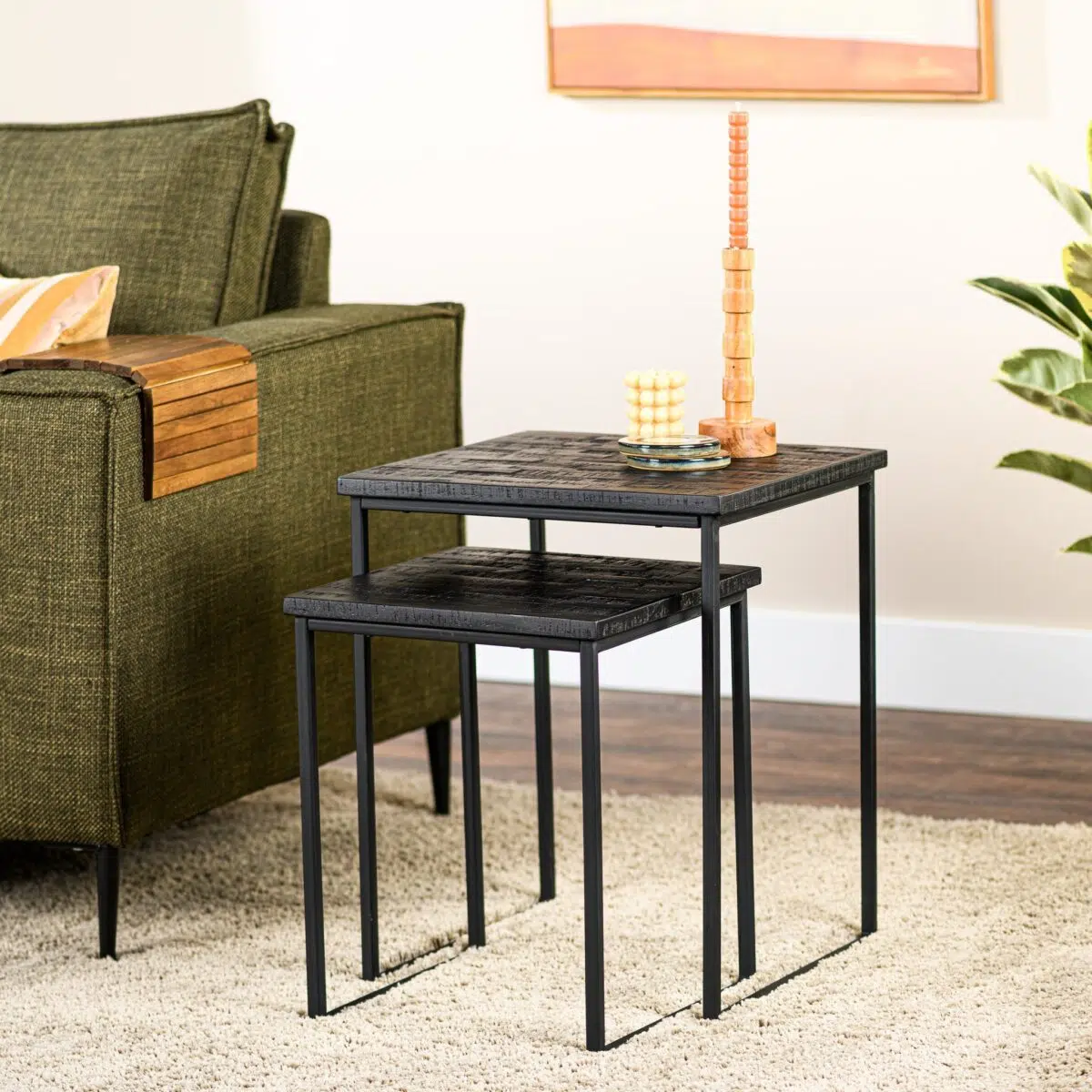 Twee moderne bijzettafels met zwarte metalen frames en houten bladen staan naast een groene bank. Een stapel houten blokken en een klein decoratief item staan op de grotere tafel. Een plant en een ingelijst kunstwerk staan op de achtergrond.
