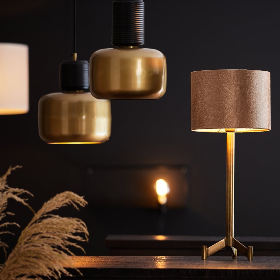 Moderne interieurscène met een houten tafellamp op de voorgrond en drie hangende hanglampen op de achtergrond. De hanglampen zijn van metaal met een donkere afwerking. Links zijn decoratieve grassen zichtbaar, allemaal tegen een donkere muur.