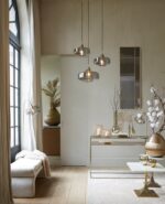 Een stijlvolle woonkamer met een minimalistisch design met hangende hanglampen, een consoletafel met kaarsen en vazen, een witte bank met een deken en een groot raam met doorschijnende gordijnen. Aardse accenten en moderne inrichting maken de ruimte compleet.