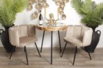 Een kleine ronde tafel met een decoratief octopus-middenstuk, geflankeerd door twee beige stoelen. De tafel bevat twee wijnglazen en een fles. Bronzen bladmuurkunst is op de achtergrond te zien, met hoge groene planten in donkere potten aan beide kanten.