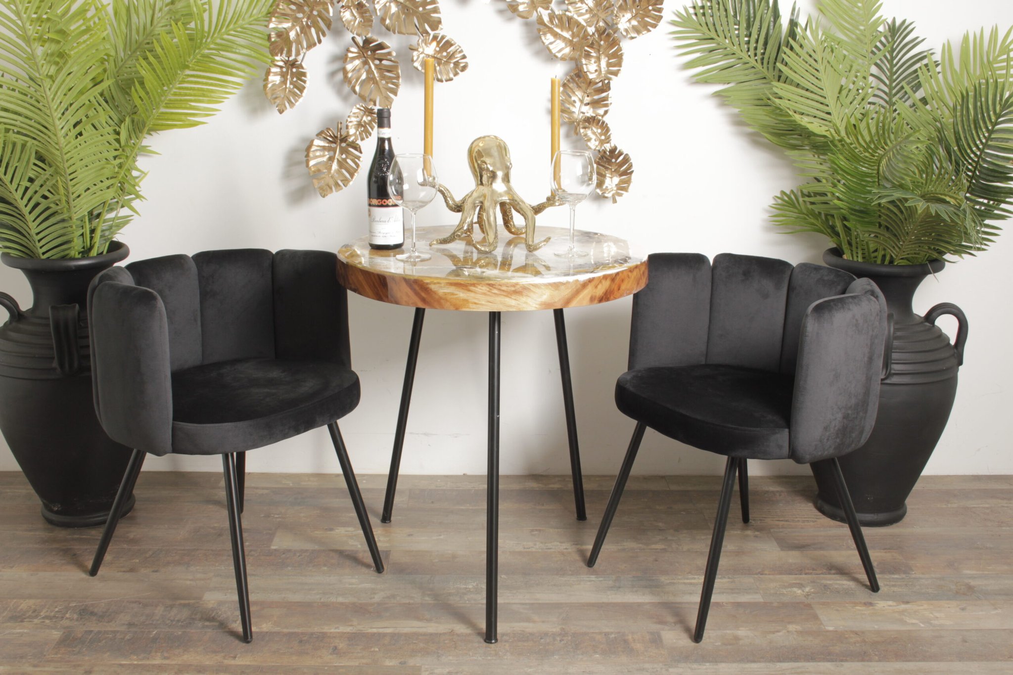Een ronde tafel met een gouden octopus als middelpunt is gedekt met wijn en glazen. Twee zwarte fluwelen stoelen omringen het. Op de achtergrond zorgen decoratieve bladgouden wandkunst en hoge potplanten voor een weelderige touch. De vloer is van hout.