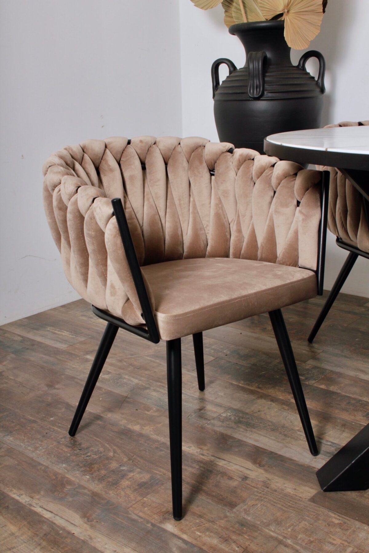 Een moderne beige fluwelen eetkamerstoel met een gewatteerde rugleuning en zwarte metalen poten staat op een houten vloer. Ernaast staat een ronde tafel. Een grote zwarte vaas met decoratieve gedroogde bladeren staat op de achtergrond.