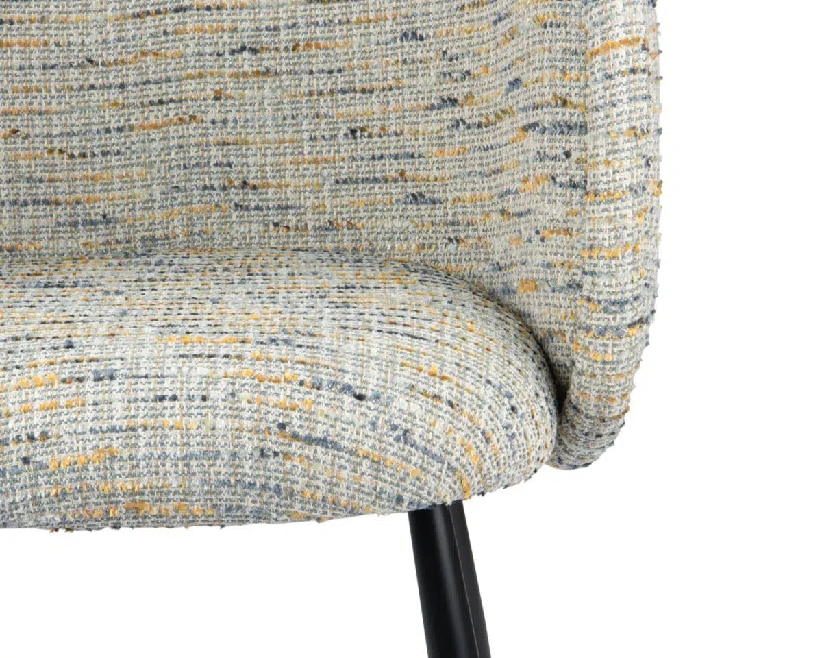 Close-up van een stoel met textuurstof met een gebogen rug en zitting in een gespikkeld patroon van crème, grijs en mosterdkleuren. De stoel heeft een zwarte metalen poot die zichtbaar is aan de onderkant.