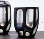 Twee grote, zwarte geometrische vazen met reflecterende, doorschijnende panelen staan op een donker houten oppervlak. De vazen hebben een hoekig, modern ontwerp met een onregelmatig patroon op hun oppervlakken.