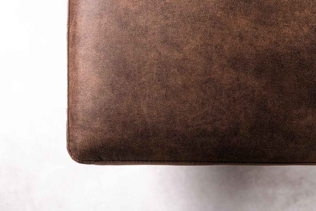 Close-up van een rechthoekig, bruin leren kussen of zithoek tegen een witte achtergrond. Het leer heeft een gladde textuur met lichte kleurvariaties, wat duidt op een versleten of vintage look.