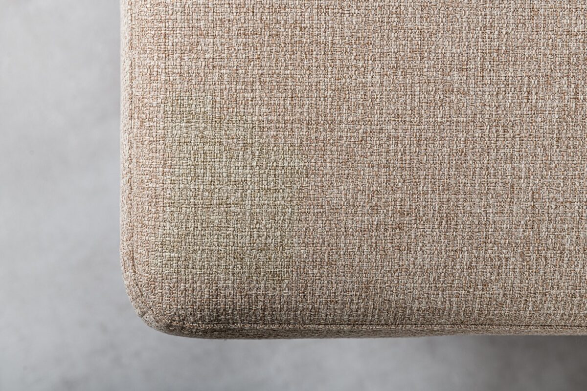Close-up van een beige stoffen hoekbank met een zichtbare vlek aan de linkerkant. De textuur van het geweven materiaal is gedetailleerd en de achtergrond is een lichtgrijs, wazig oppervlak.